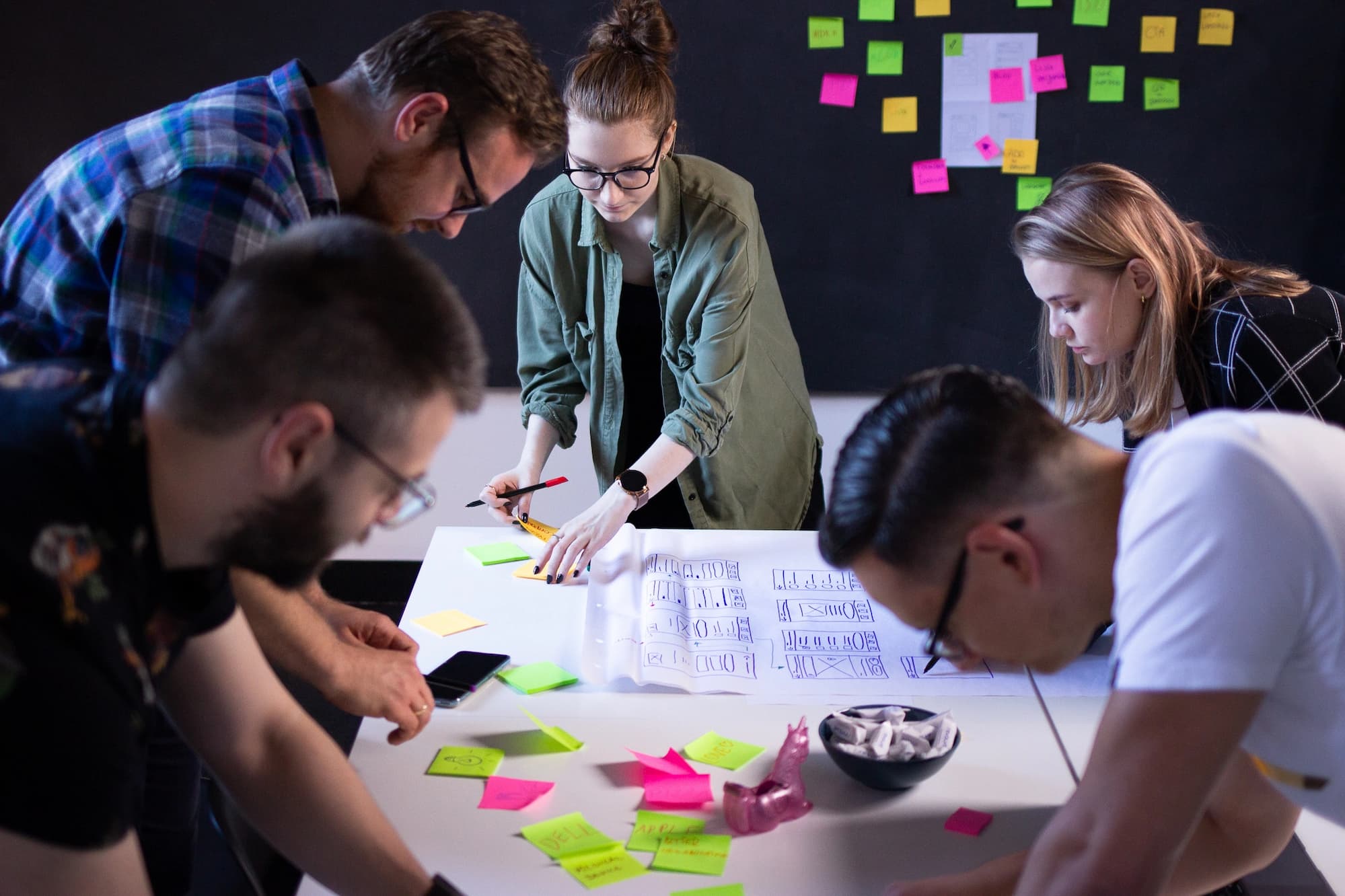 Design workshops – five people standing over a table, with many sticky notes around and one woman drawing a user interface on a big sheet of paper