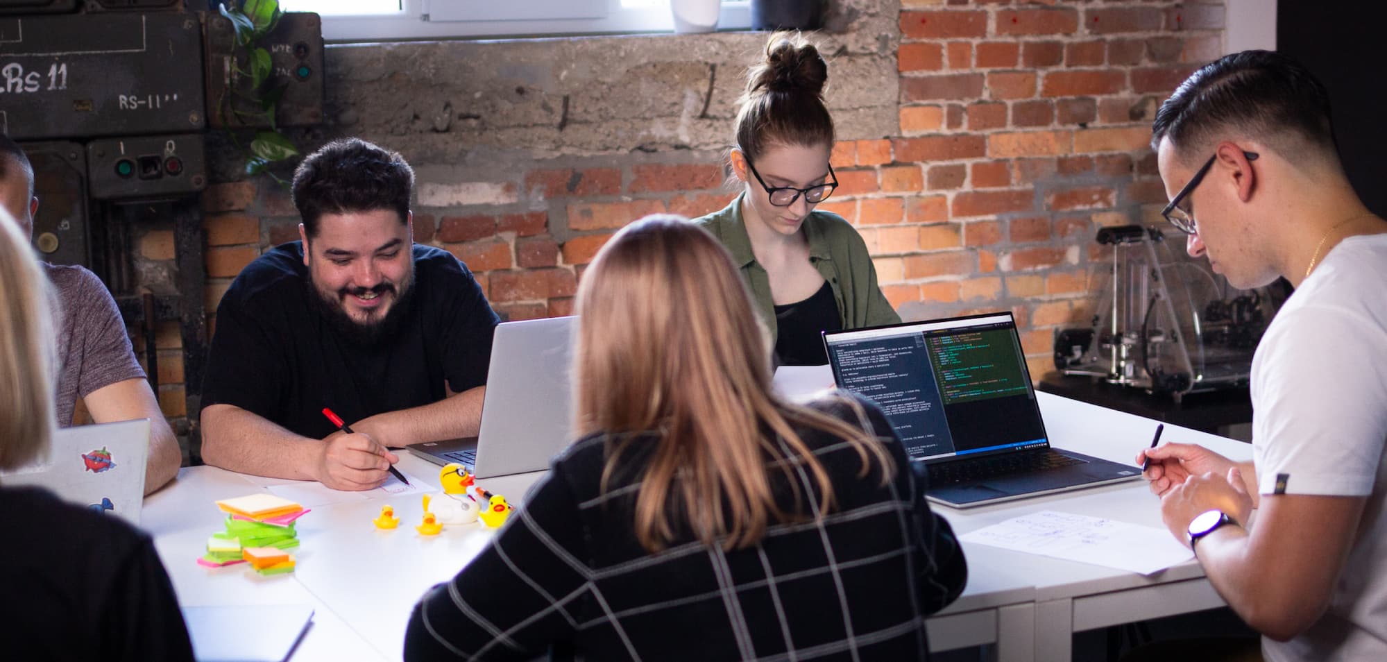 A group of people sitting at a long table with laptops and some rubber ducks on it, writing on A4 sheets of paper during a project planning meeting at an office with red brick wall
