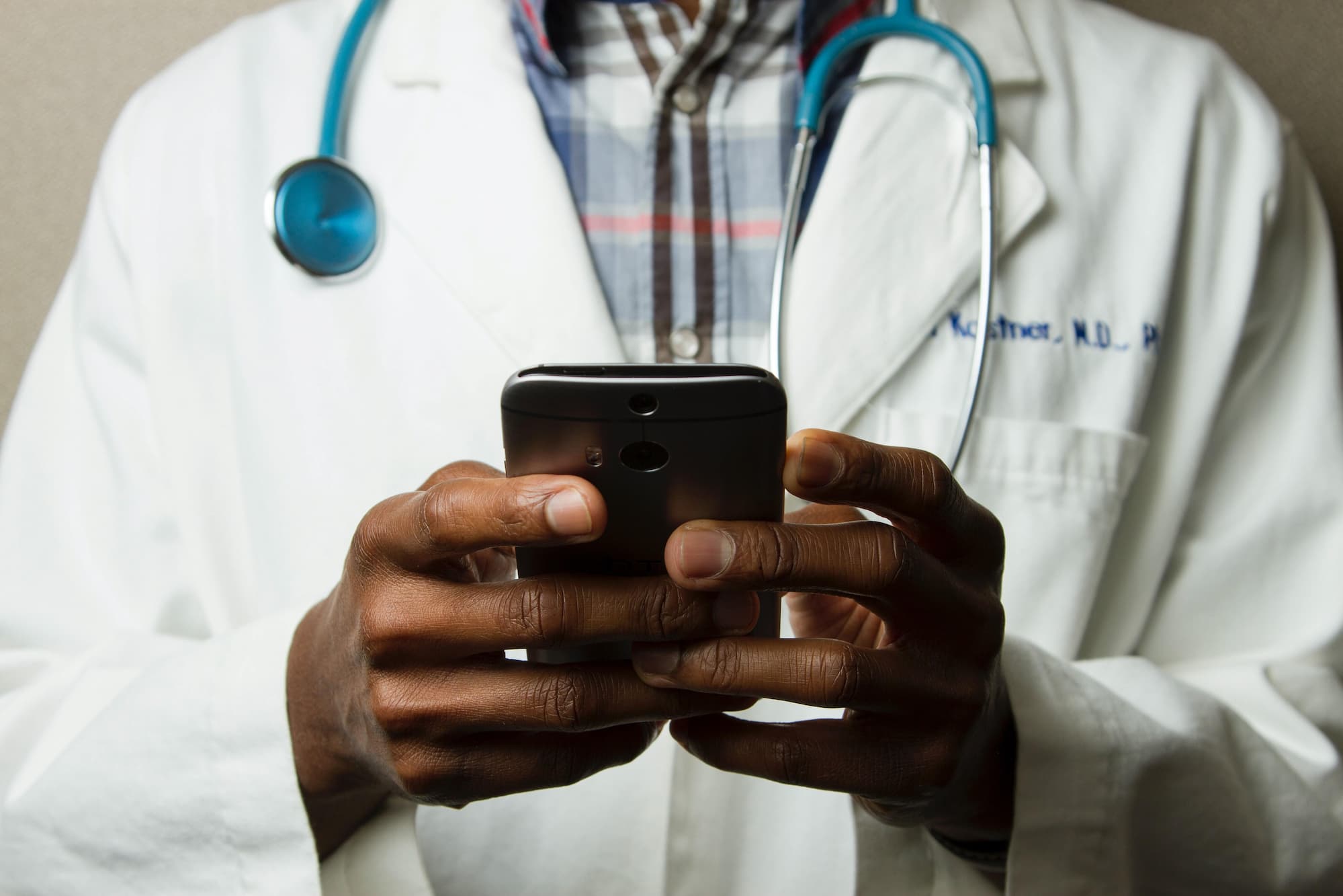 Hands of a doctor in a while overall, with a stethoscope around his neck, holding a smartphone